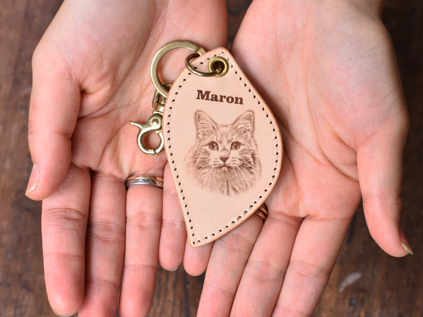 ペットレザー彫刻キーホルダーリーフ型を両手のひらの上に乗せている写真。レザーキーホルダーの中心に猫の顔写真があり、上にMaronと書いている。