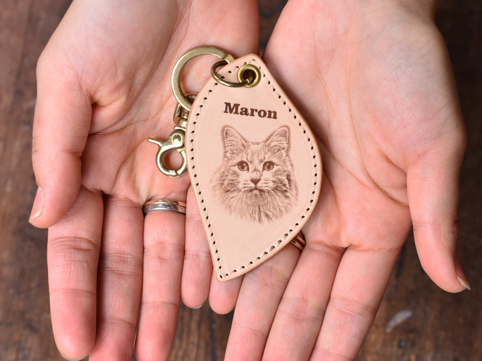 ペットレザー彫刻キーホルダーリーフ型を両手のひらの上に乗せている写真。レザーキーホルダーの中心に猫の顔写真があり、上にMaronと書いている。